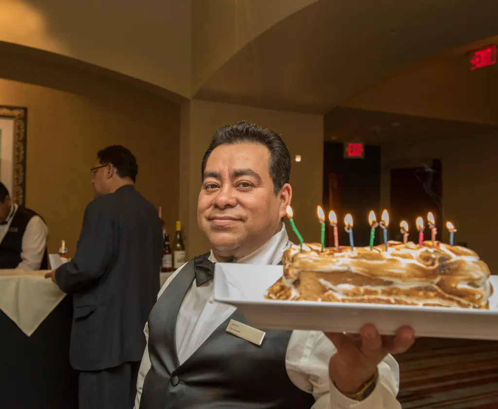 A man in a black vest and bow tie holds a long cake with toasted meringue and a row of lit candles.