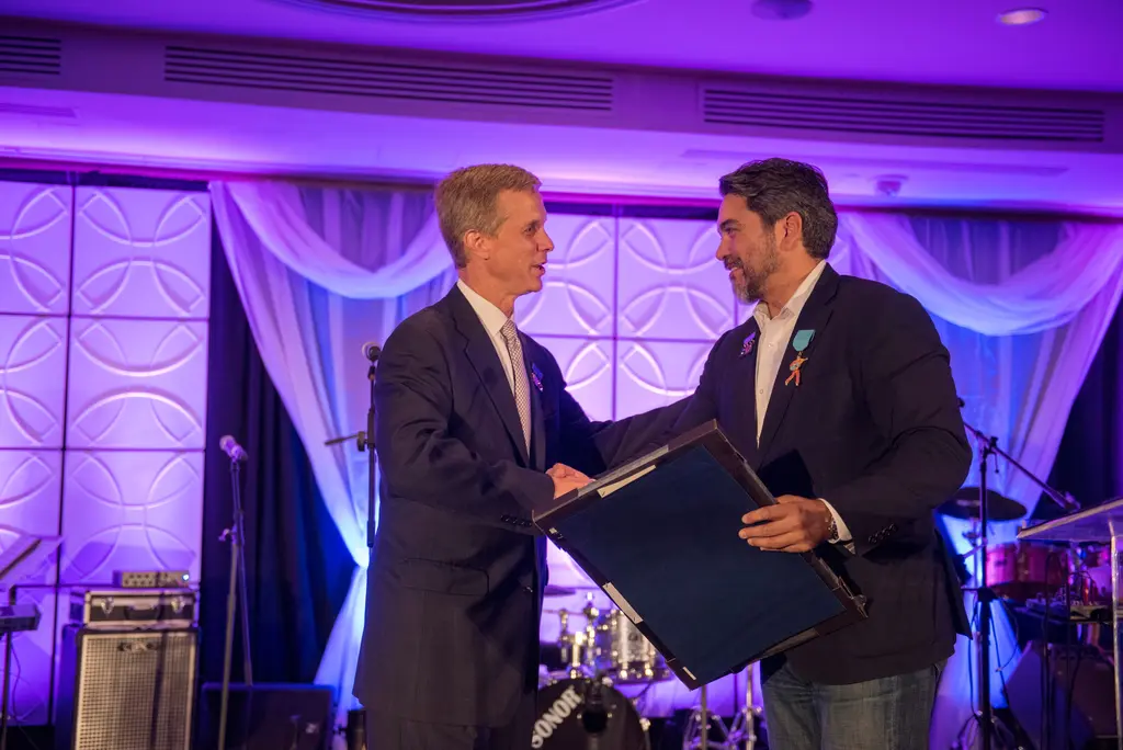 Two men in dark jackets shake hands in front of purple-lit drapes while one holds a framed certificate.