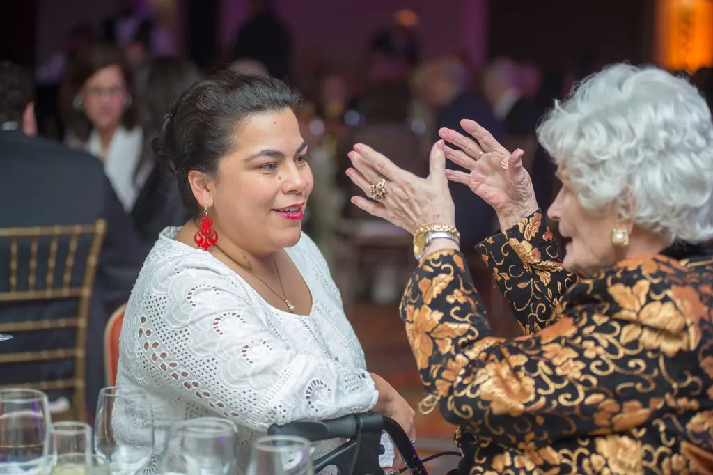 A woman in a white lace top and red earrings listens as an older woman in a black and gold jacket gestures with both hands.