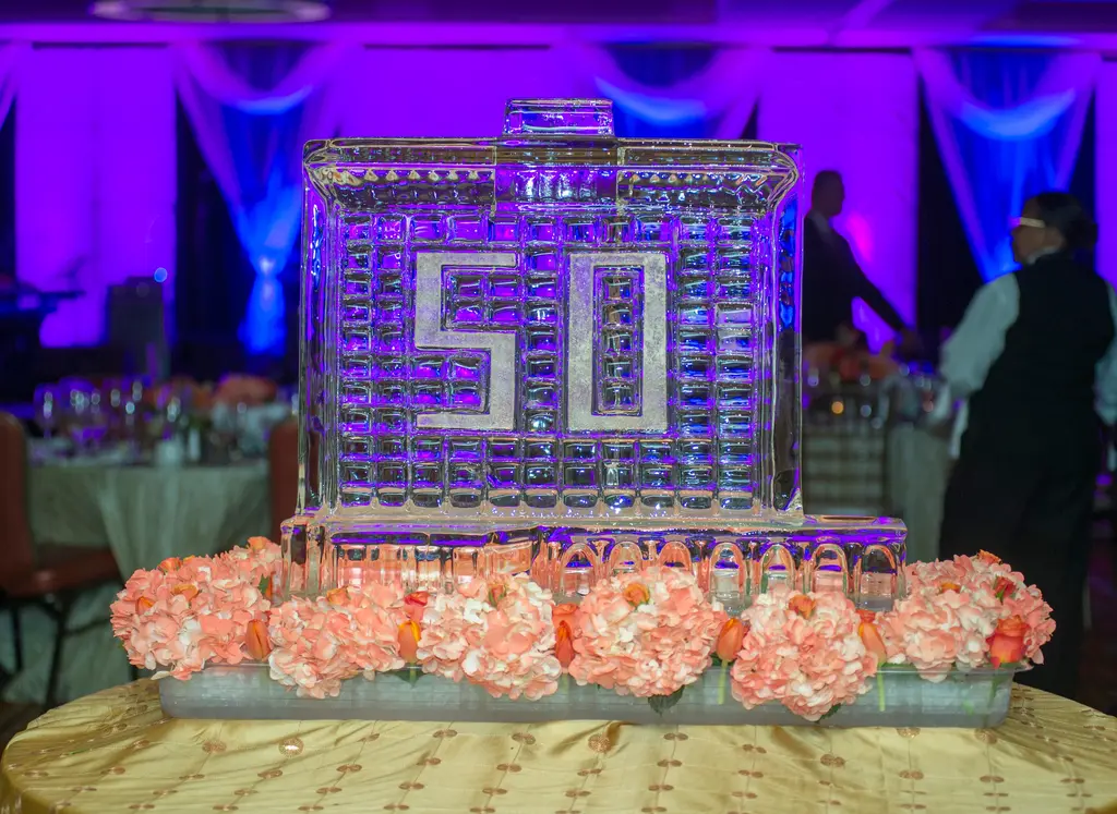 A clear ice sculpture shaped like a high-rise hotel with the number 50 carved into its face, lit purple and set on peach flowers on a gold tablecloth.