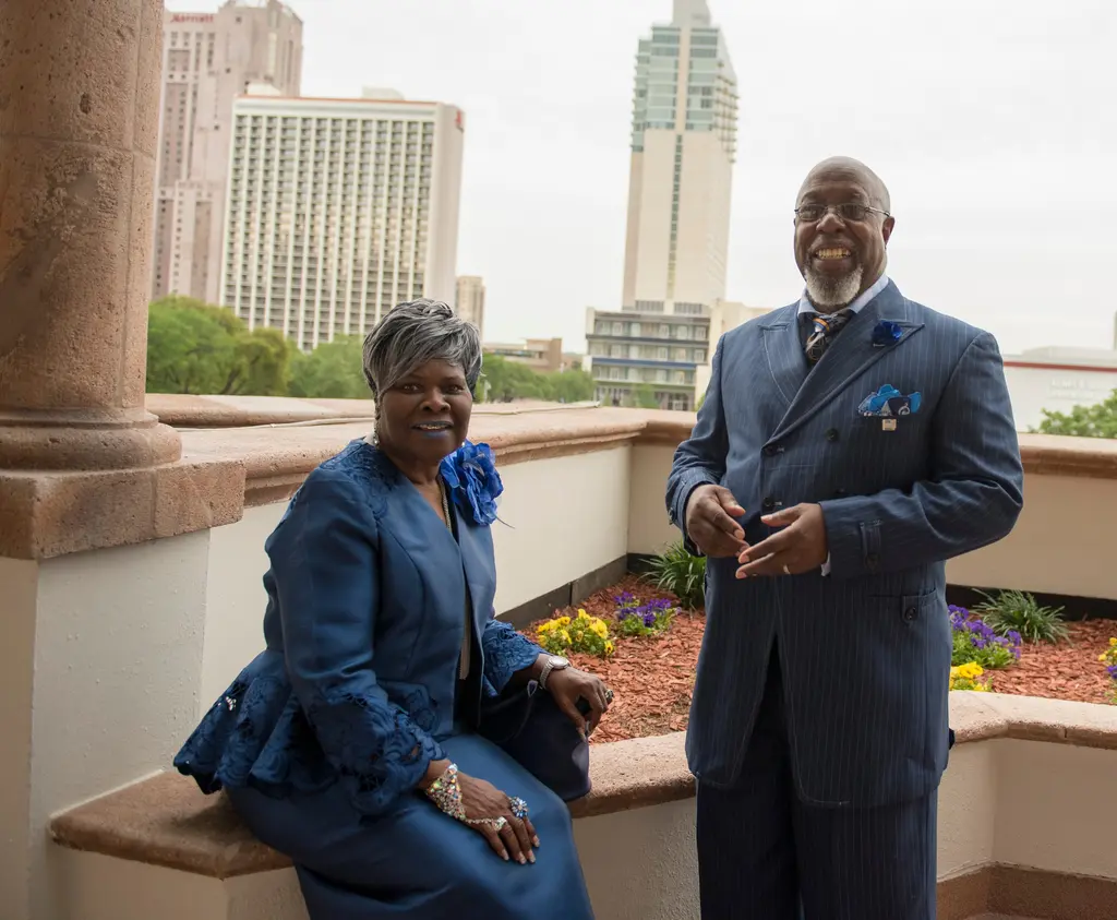 A smiling woman in a blue suit with a flower corsage sits on a terrace ledge beside a laughing man in a blue pinstripe suit, with downtown high-rises behind them.