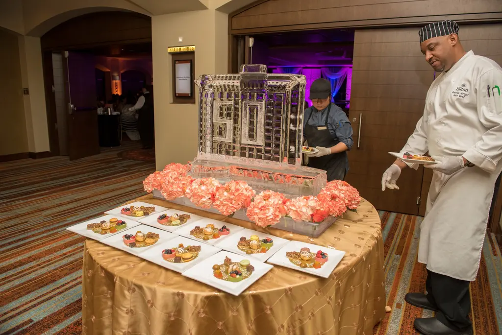 A chef in a white Hilton jacket and striped cap and a cook in a black cap carry dessert plates beside an ice sculpture ringed with small plated desserts on a gold table.