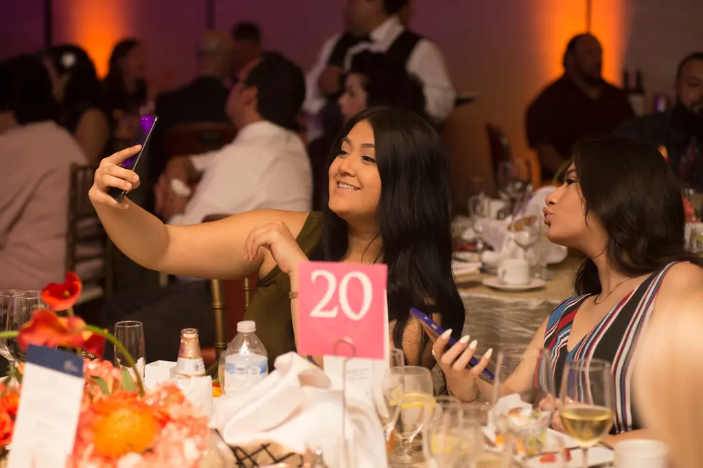A young woman holds up a phone for a selfie while her friend puckers her lips beside her, behind a pink table card with the number 20.
