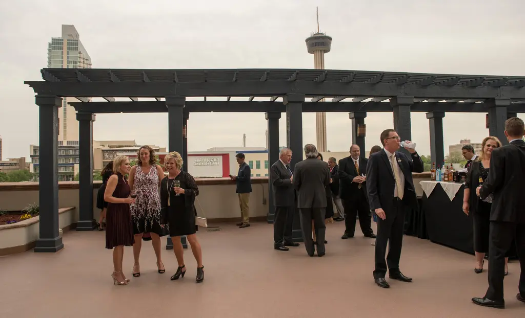 Guests in cocktail attire stand and talk on a wide rooftop terrace under a dark wooden pergola, with the Tower of the Americas and downtown buildings in the background.
