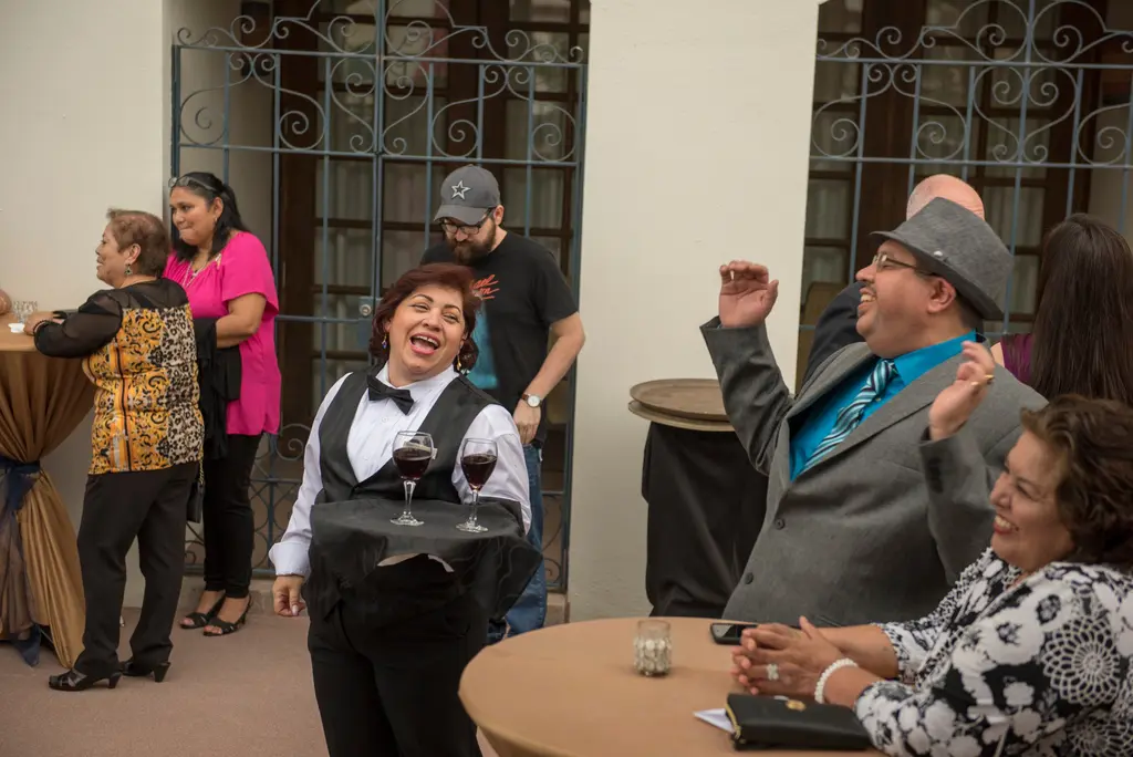 A smiling server in a black vest and bow tie holds a tray with two glasses of red wine while a man in a gray hat raises his hands and a seated woman laughs.
