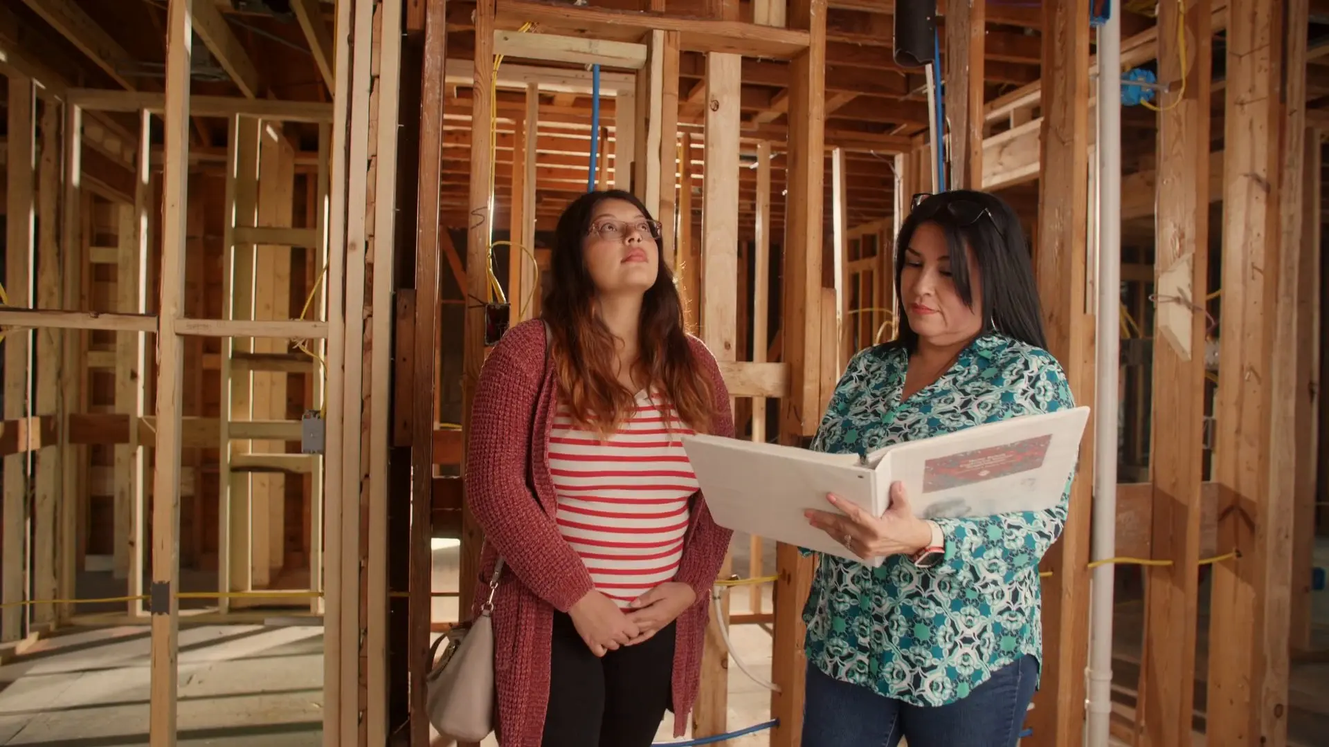 A woman reads from an open binder as another woman looks up at the exposed wood framing inside a house.