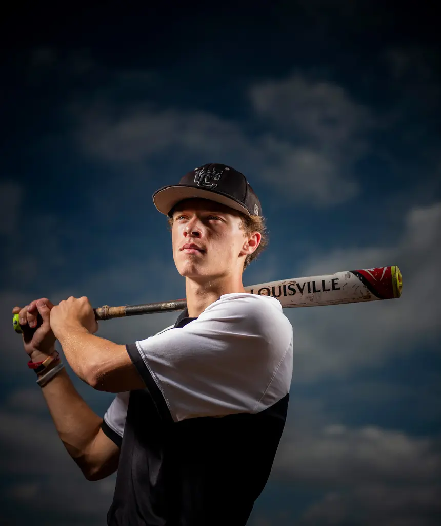 A young baseball player in a black cap rests a Louisville bat on his shoulder under a dark, cloudy sky.