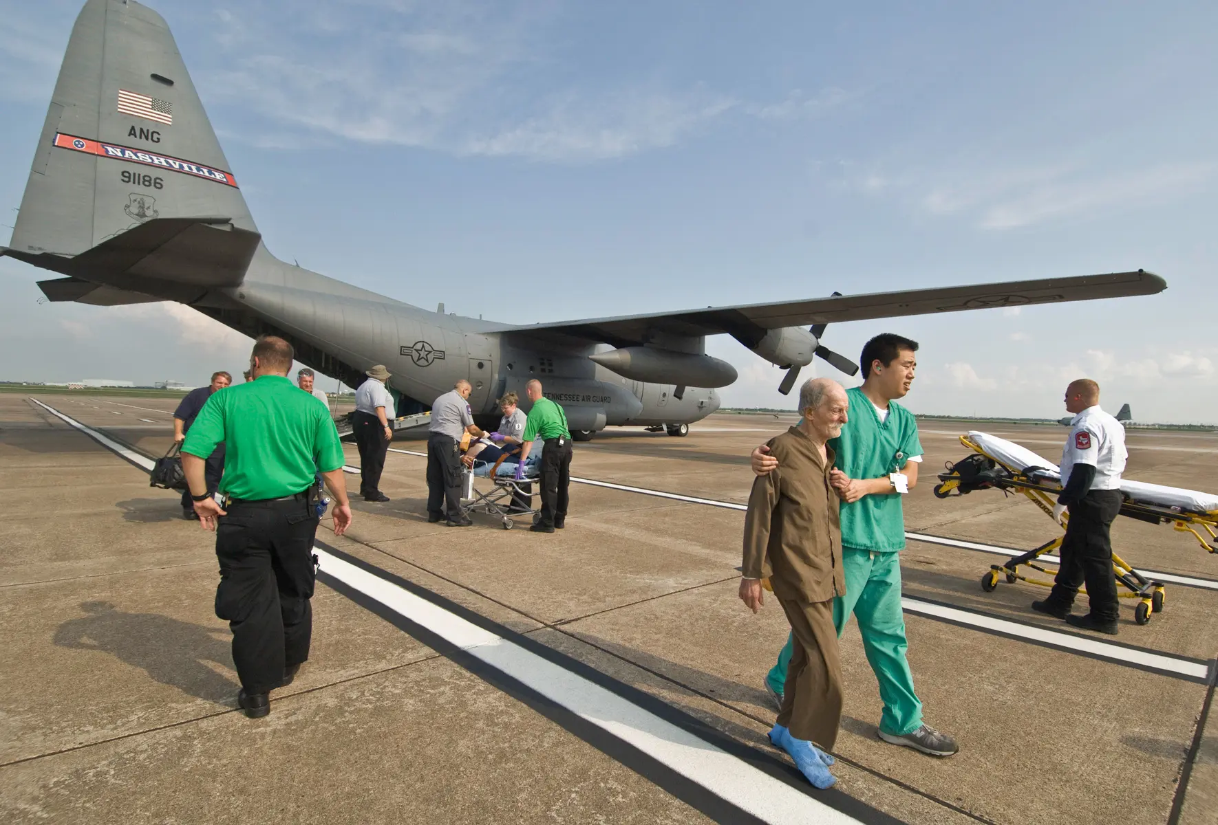 A man in green scrubs walks a barefoot elderly man in brown pajamas across a tarmac away from a gray military cargo plane, with stretchers and crews behind them.