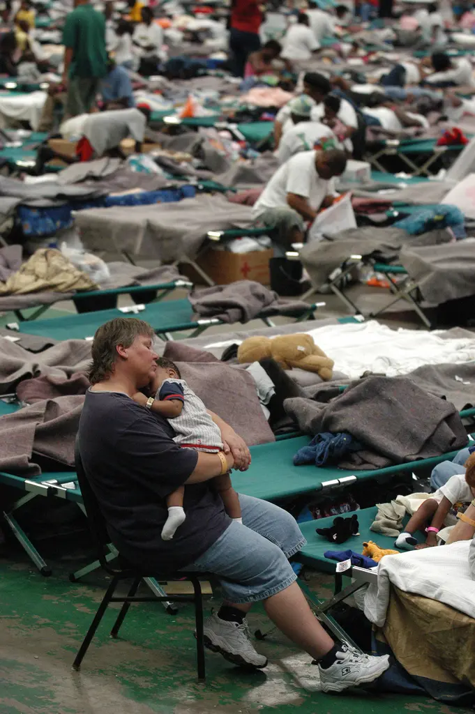 A volunteer sleeps sitting up in a chair, holding a sleeping baby, among rows of green cots on the floor of the Reliant Astrodome.