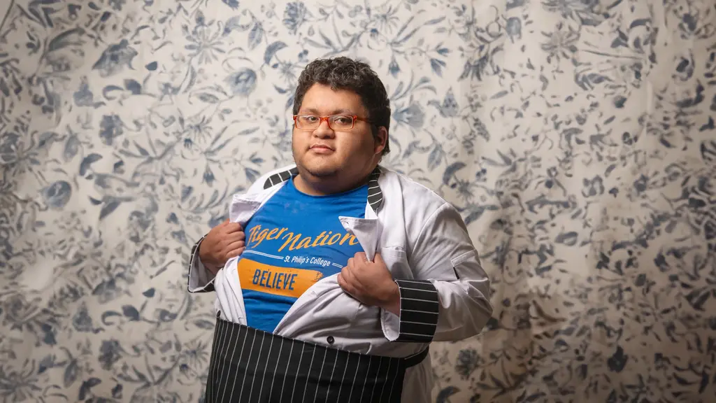 Jacob Gutierrez, in a chef coat and striped apron, pulls his coat open to show a blue Tiger Nation shirt from St. Philip’s College against floral wallpaper.