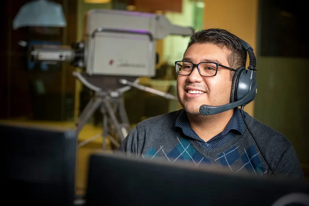 Joel Valadez in a headset smiles at a control desk beside a vintage television camera