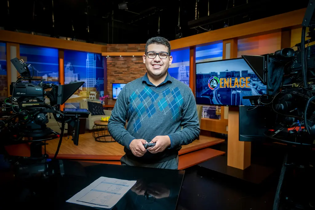Joel Valadez stands on a television news set among studio cameras and monitors