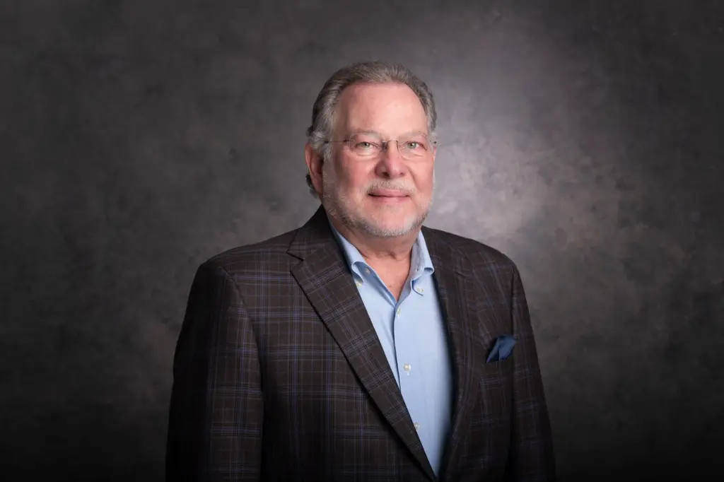 The CEO of Joeris General Contractors, in glasses, a plaid sport coat and a light blue shirt, against a gray studio backdrop.