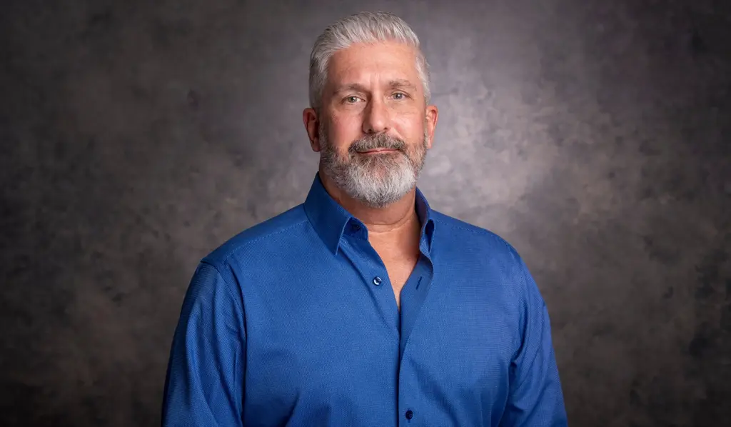 A Joeris General Contractors executive with silver hair and a gray beard, in a blue button-down shirt, against a gray studio backdrop.