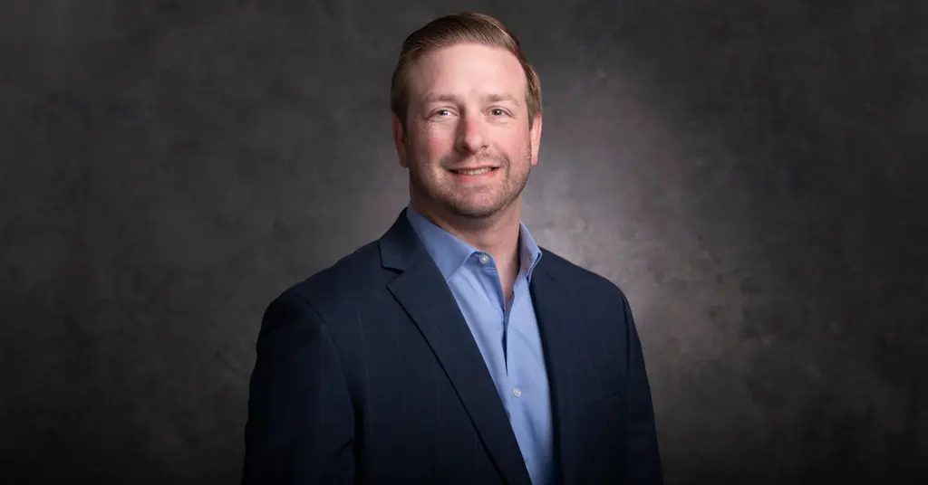 A smiling Joeris executive in a navy windowpane blazer and light blue shirt, against a gray studio backdrop.