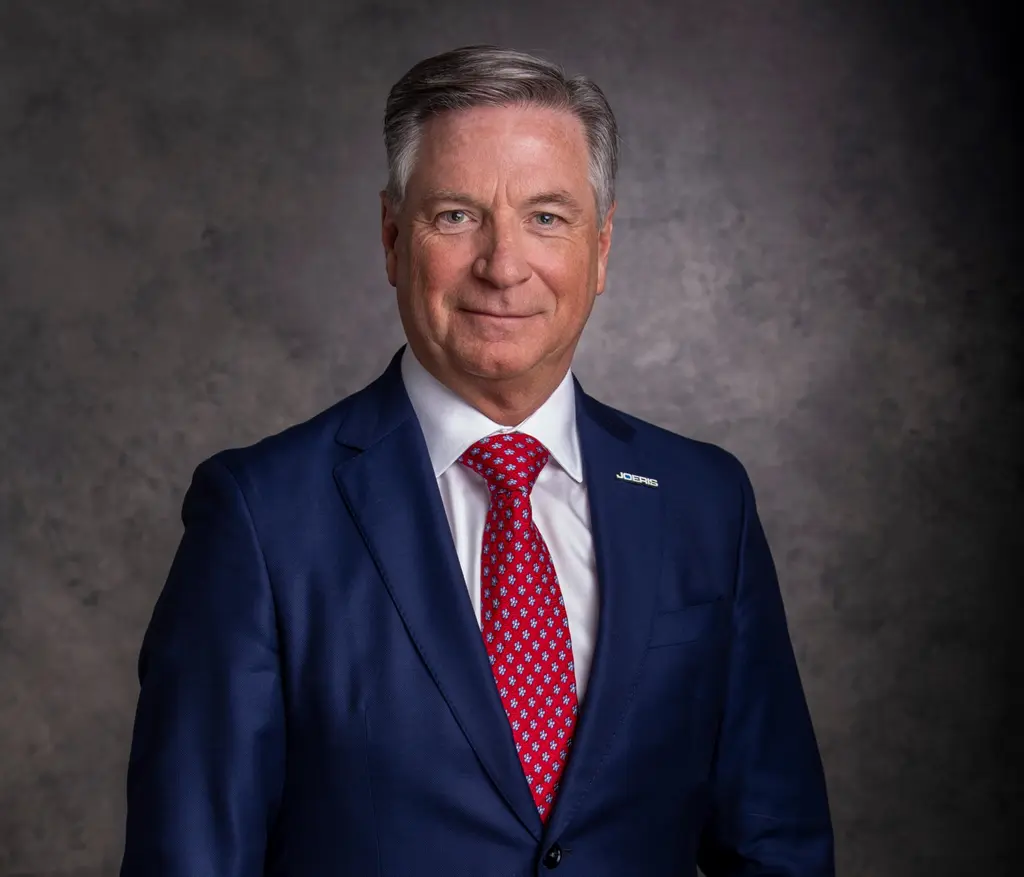 A Joeris executive in a navy suit, a red patterned tie and a Joeris lapel pin, against a gray studio backdrop.