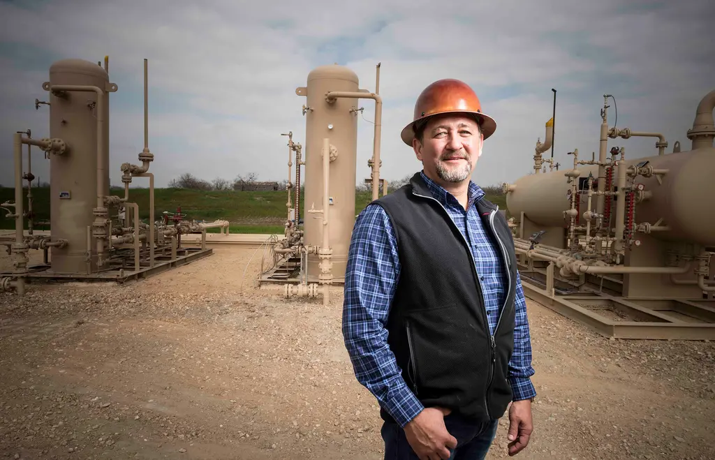Joseph Dorn in a copper hard hat, plaid shirt and black vest stands among oil and gas separators and tanks