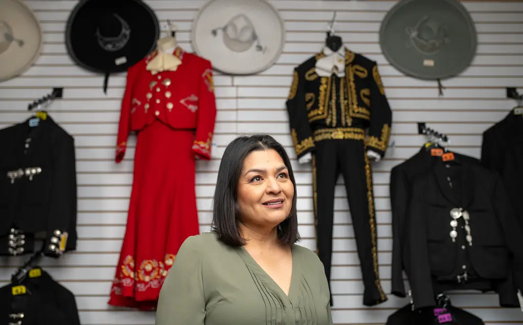 Josie Benavidez in a green blouse stands in front of charro suits and sombreros at her shop