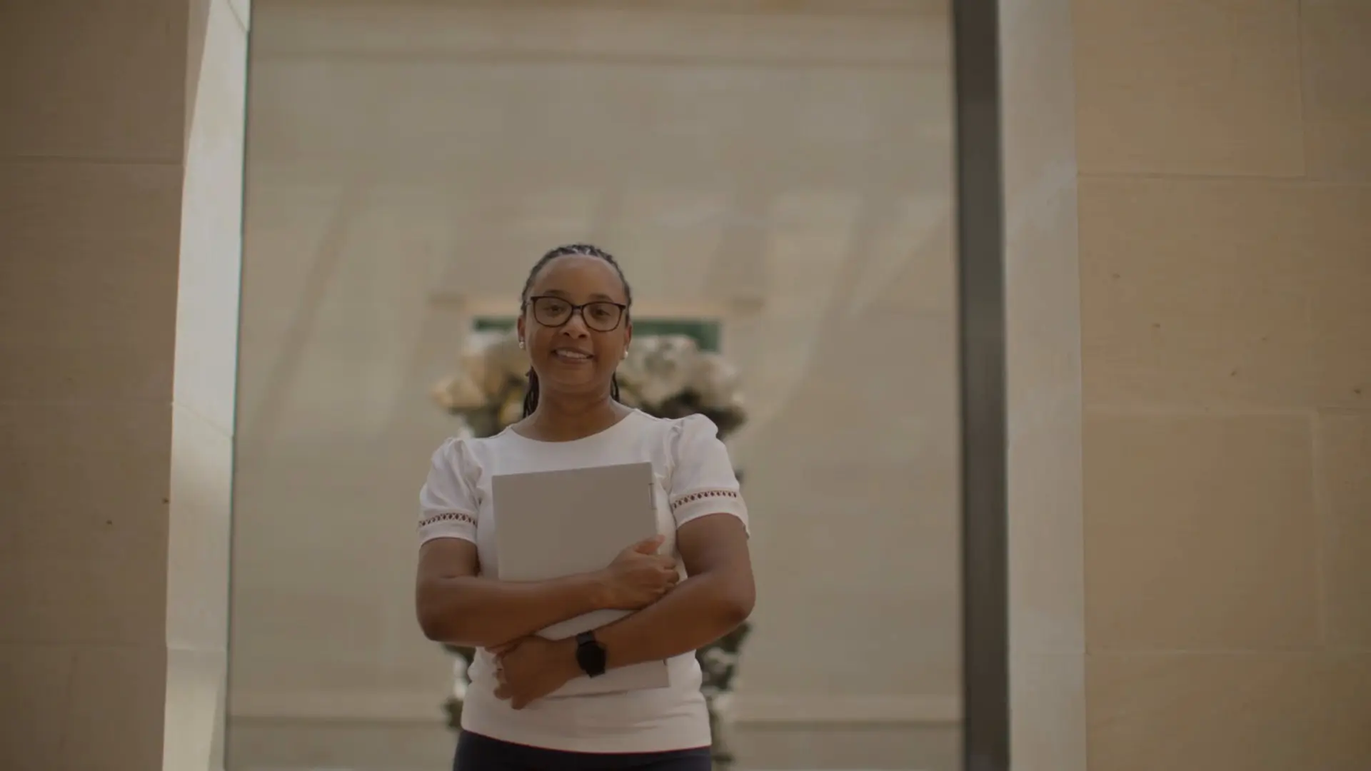 Kayla Guillory smiles in glasses and a white top, hugging a silver laptop to her chest in a cream stone hallway with flowers behind her.