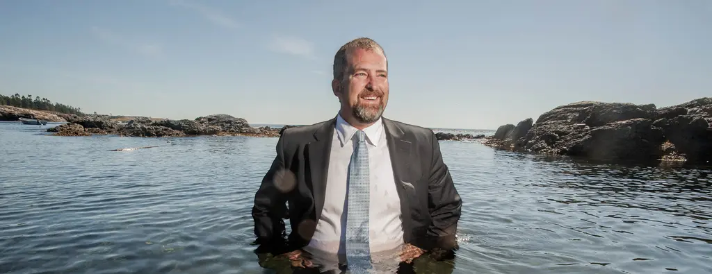 Kevin Ranker, who represented the San Juan Islands in the Washington state Senate, stands in a suit and tie, waist-deep in the water off a rocky shoreline.