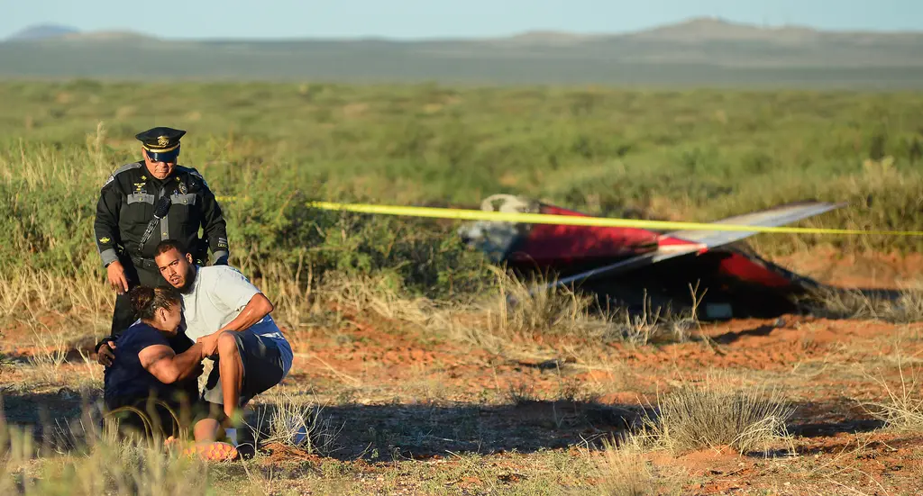 An officer stands over a woman and a man crouched in the desert brush near crime scene tape, with the wreckage of a small plane behind them.