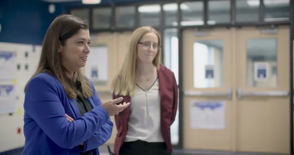 Miriam Martinez, in a royal blue blazer, gestures as she talks beside Jessica Kane, in a burgundy blazer and glasses, in a school hallway with double doors behind them.