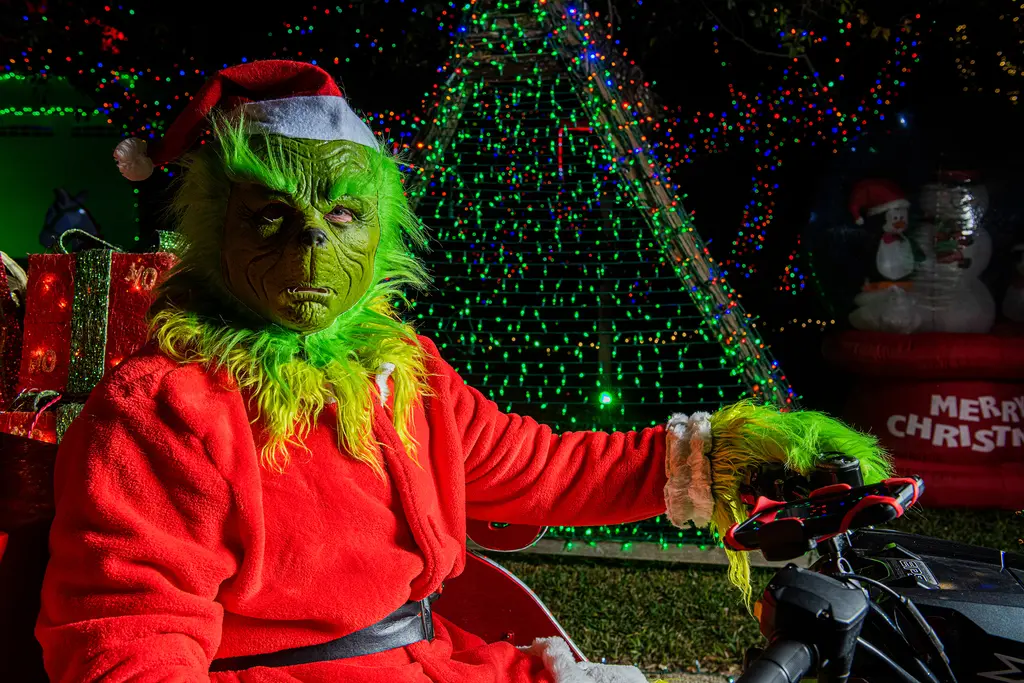 A man dressed as the Grinch sits on a light-covered ATV sleigh in front of a tree wrapped in green lights and a Merry Christmas inflatable.