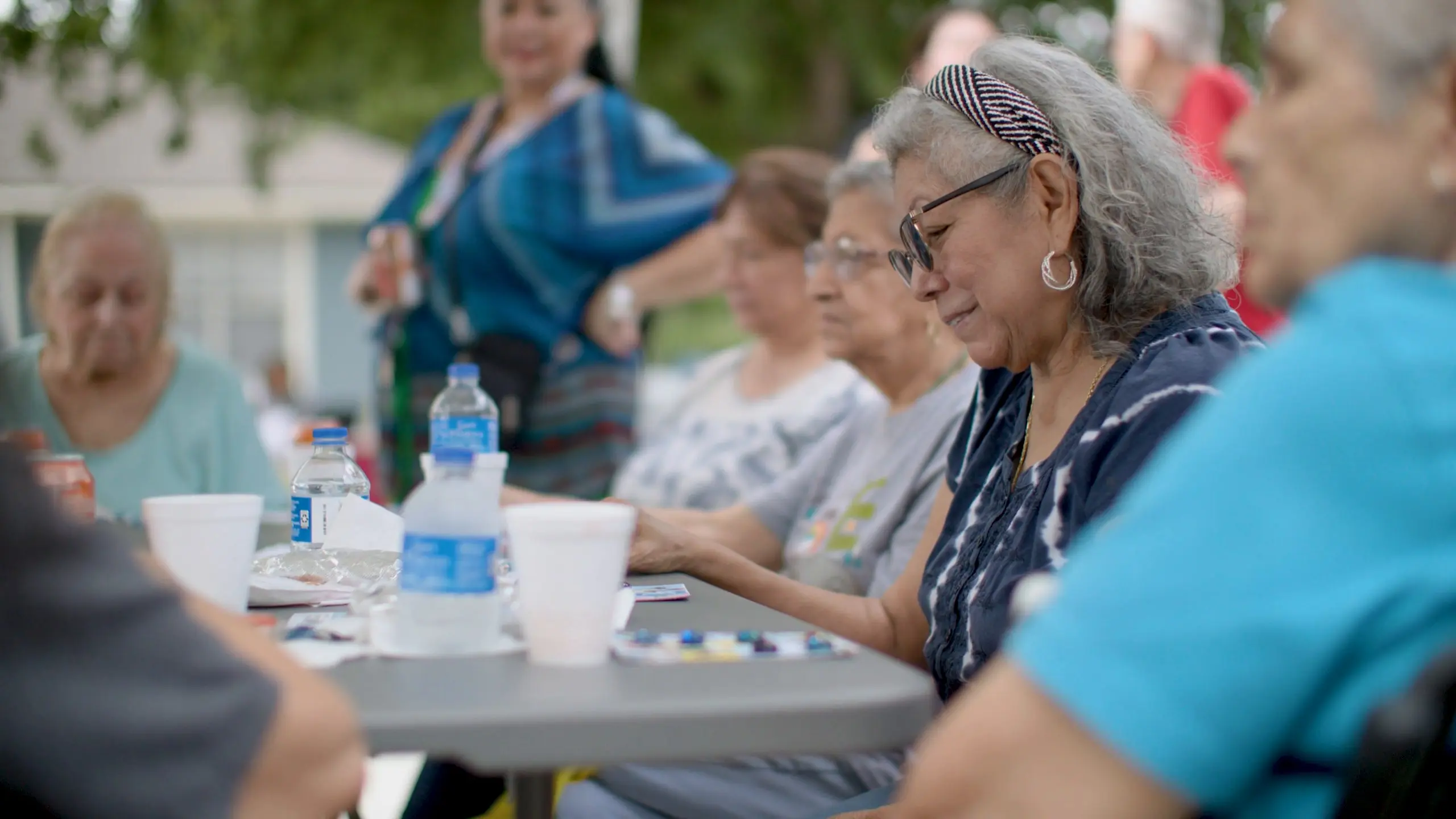 A smiling older woman with gray hair, a striped headband and glasses plays a table game with marble markers among other women at an outdoor gathering.