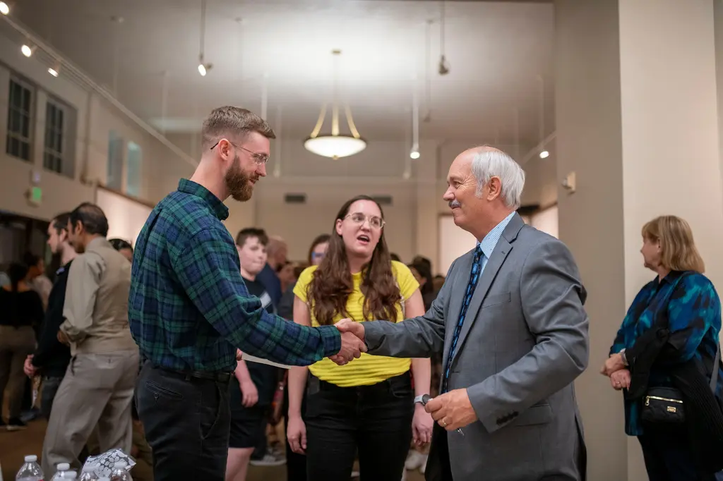 A bearded man in a plaid shirt shakes hands with a white-haired man in a gray suit as a young woman in yellow looks on.