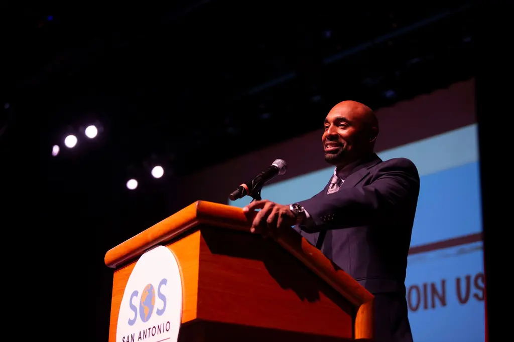A bald man in a dark suit smiles while speaking into a microphone at a wooden podium with the SOS San Antonio logo.