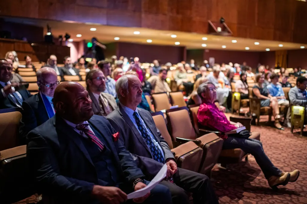Guests in suits sit in the front row of the theater holding programs, with more rows of people behind them.
