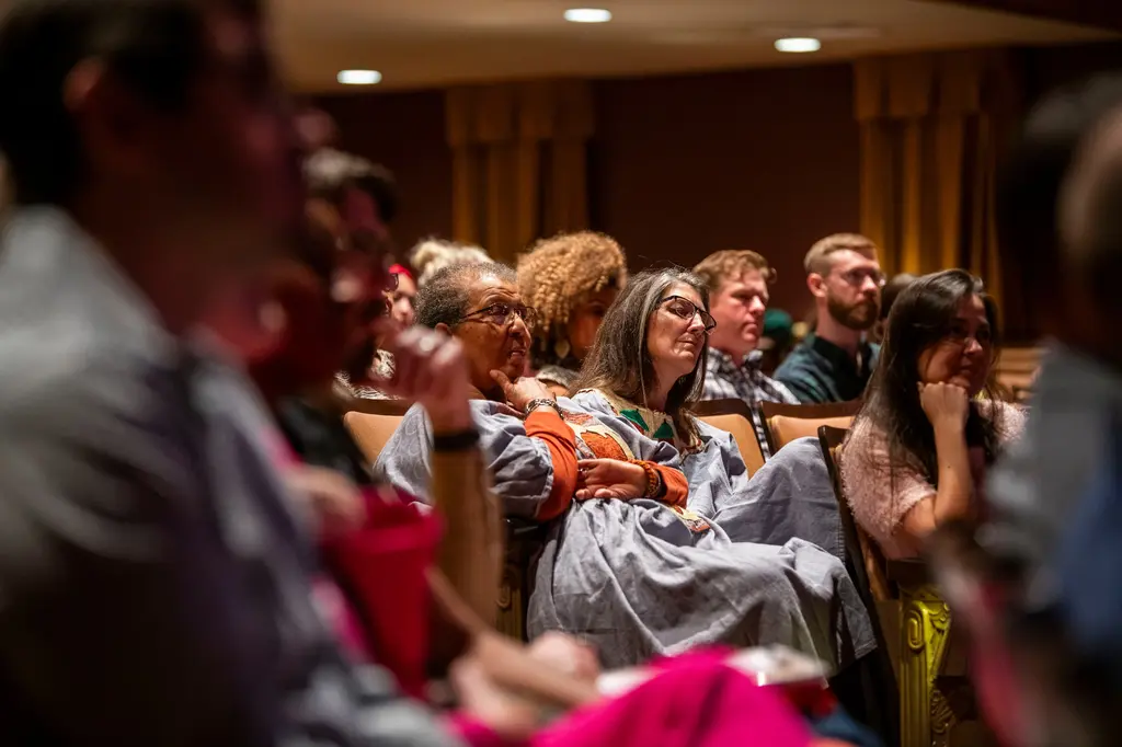 Audience members, some in African print clothing, sit in theater seats listening intently.