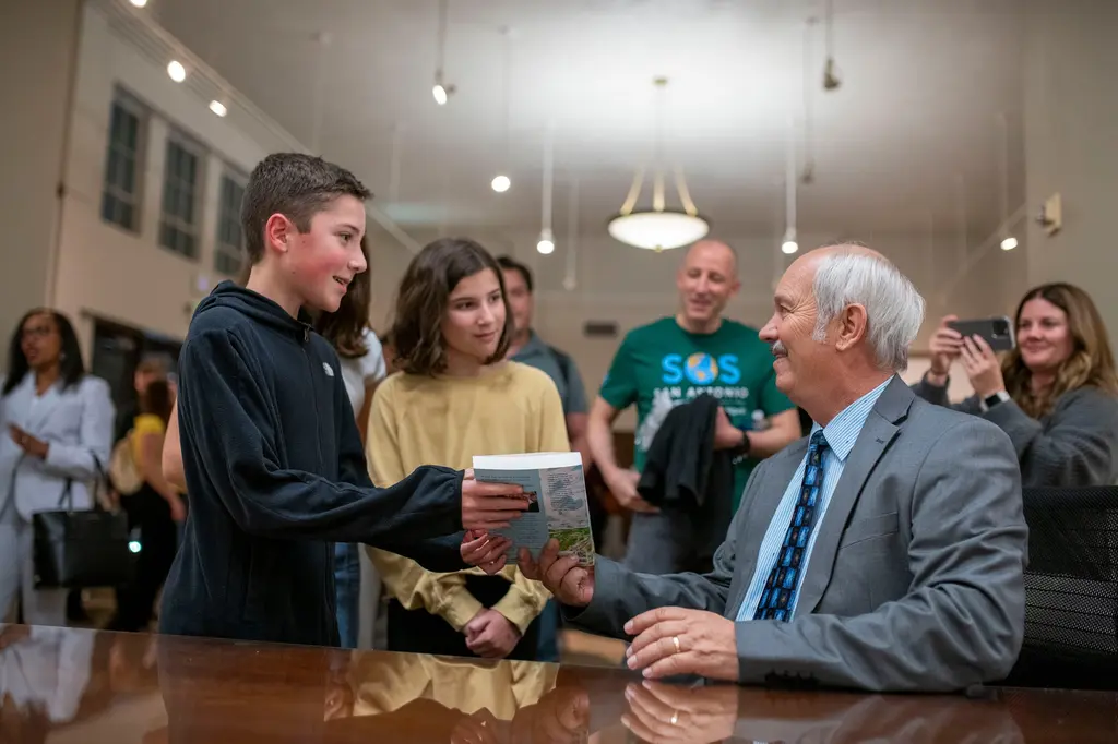 A boy in a dark hoodie hands a book across a table to a smiling white-haired man seated behind it.