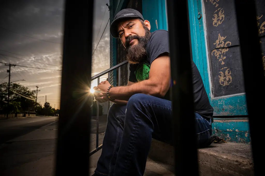Marco Cervantes sits on a stoop behind iron bars as the setting sun flares down the street