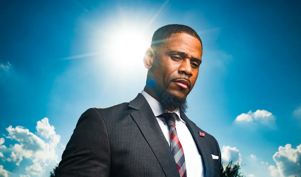Matthew Boykin in a dark suit and striped tie stands against a blue sky with the sun behind him
