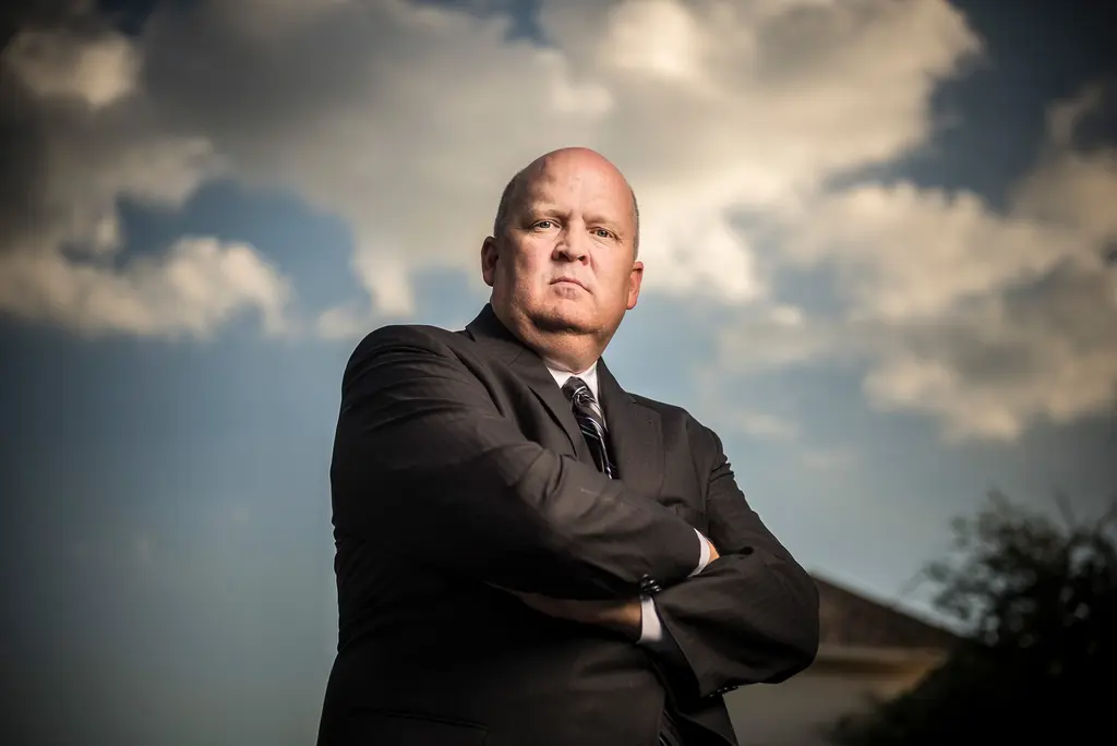 Mikal Watts in a dark suit stands with his arms crossed beneath a dramatic cloudy sky