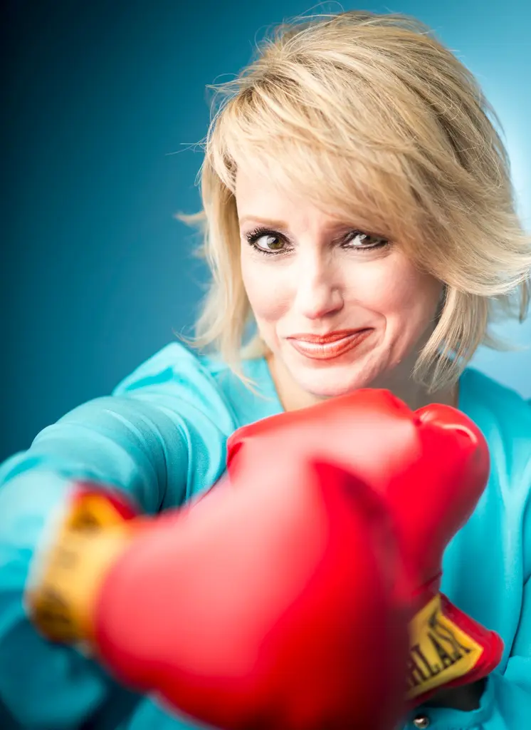 Nancy Victor, in a turquoise blouse, smiles behind a red boxing glove thrown toward the camera, against a blue backdrop.