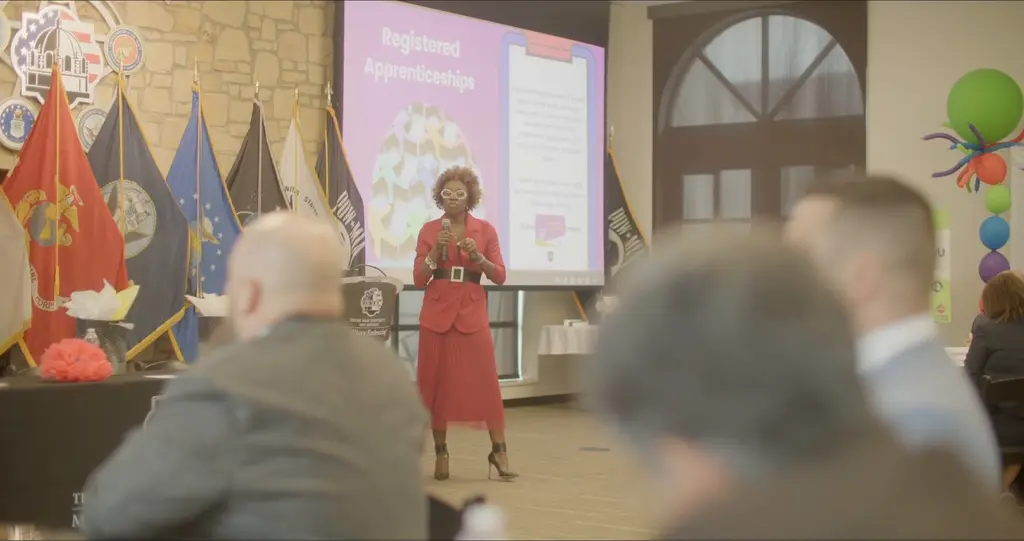 Keynote speaker Joy Jamerson, in a red suit and white glasses, speaks on stage in front of military flags and a Registered Apprenticeships slide.