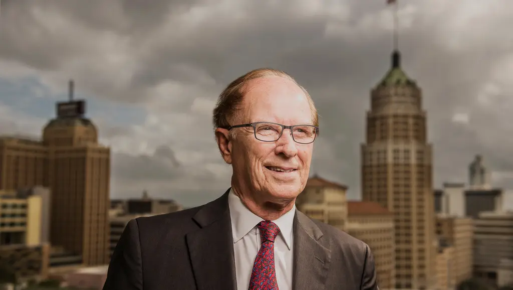 Nelson Wolff in a suit and red tie smiles with the historic towers of downtown San Antonio behind him