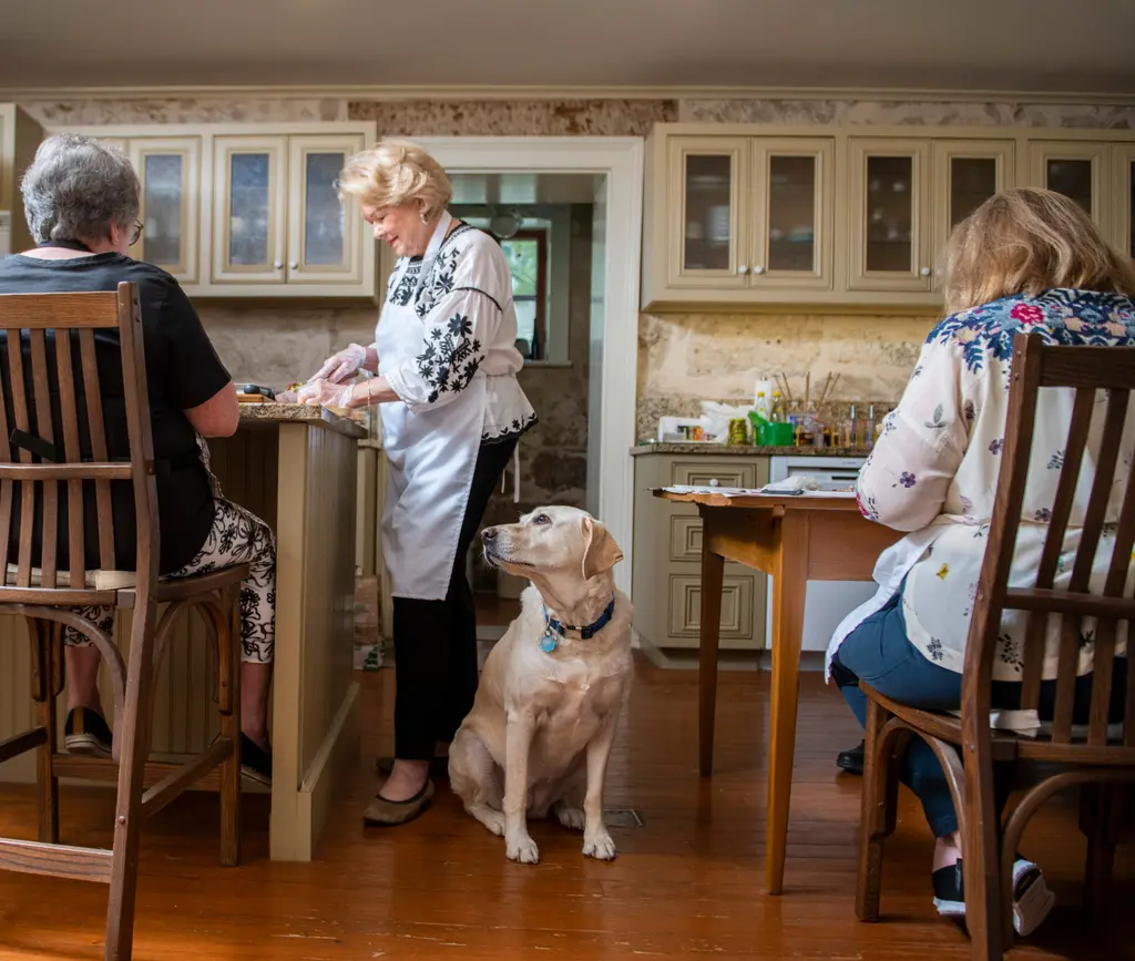 Nikki, a yellow Lab, sitting in a kitchen as women gather at a counter to prepare herring salad