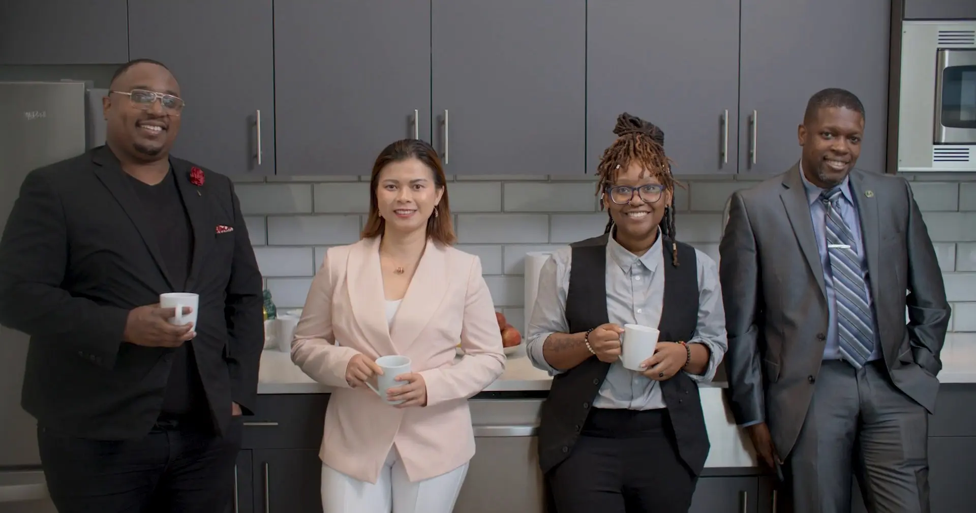 Four people in business clothes smile in an office kitchen, three of them holding white mugs.
