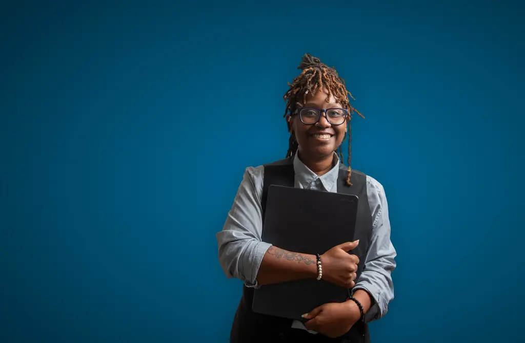 Brittany Holmes, in a gray vest, holds a black folder against a blue backdrop.