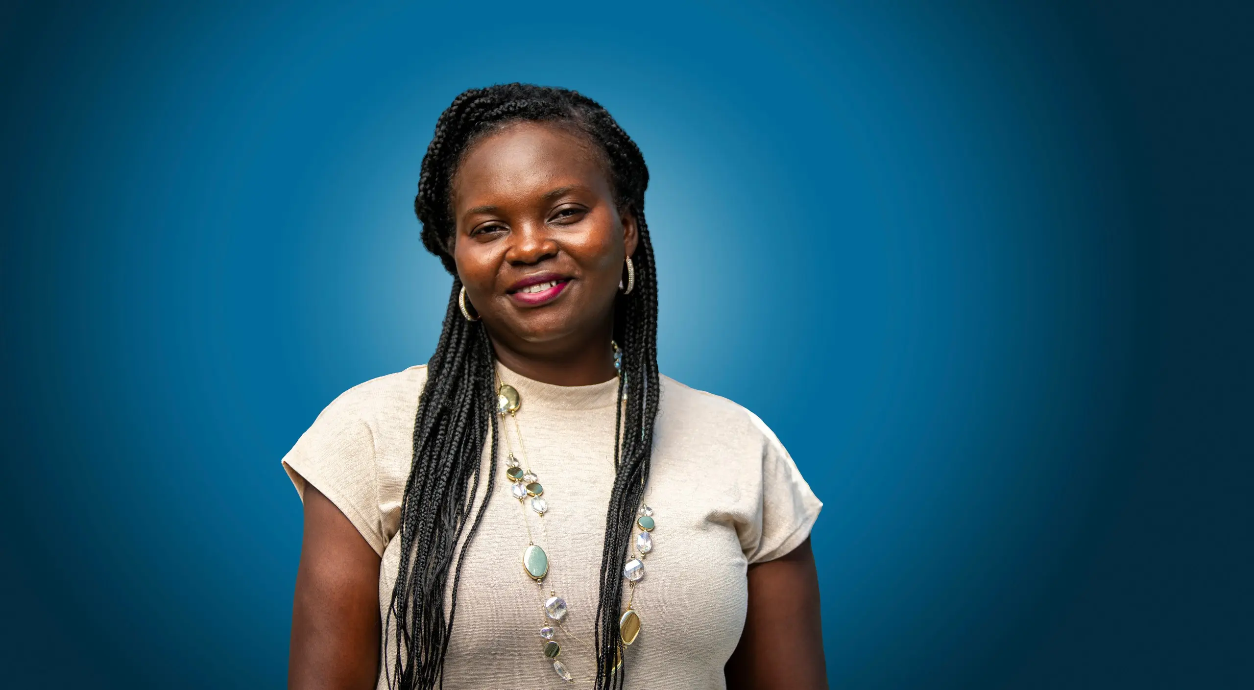 Patience Gyakari, with long braids and a layered necklace, smiles against a blue backdrop.