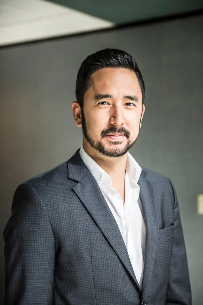 A man with a trimmed beard, in a gray checked suit and an open-collar white shirt, beside a dark office wall.