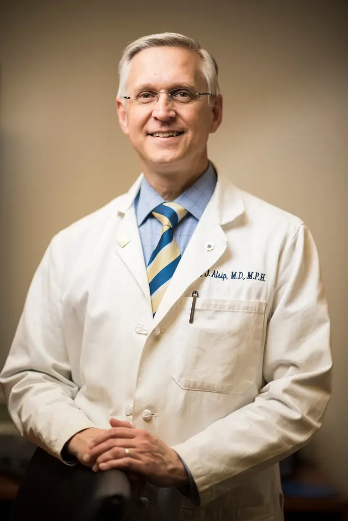 A smiling physician in glasses, a striped tie and a white lab coat embroidered with his name, his hands resting on a chair.