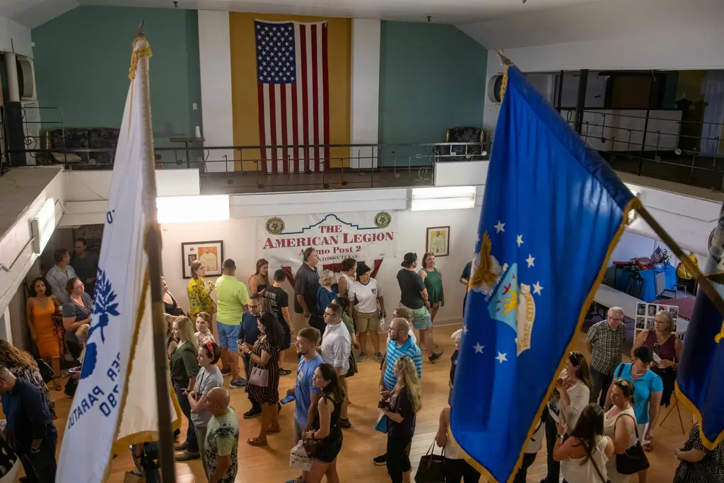 Seen past a white flag and a blue U.S. Air Force flag, a crowd mingles in a hall with a balcony, an American flag on the far wall and a banner reading The American Legion Alamo Post 2, San Antonio, Texas.