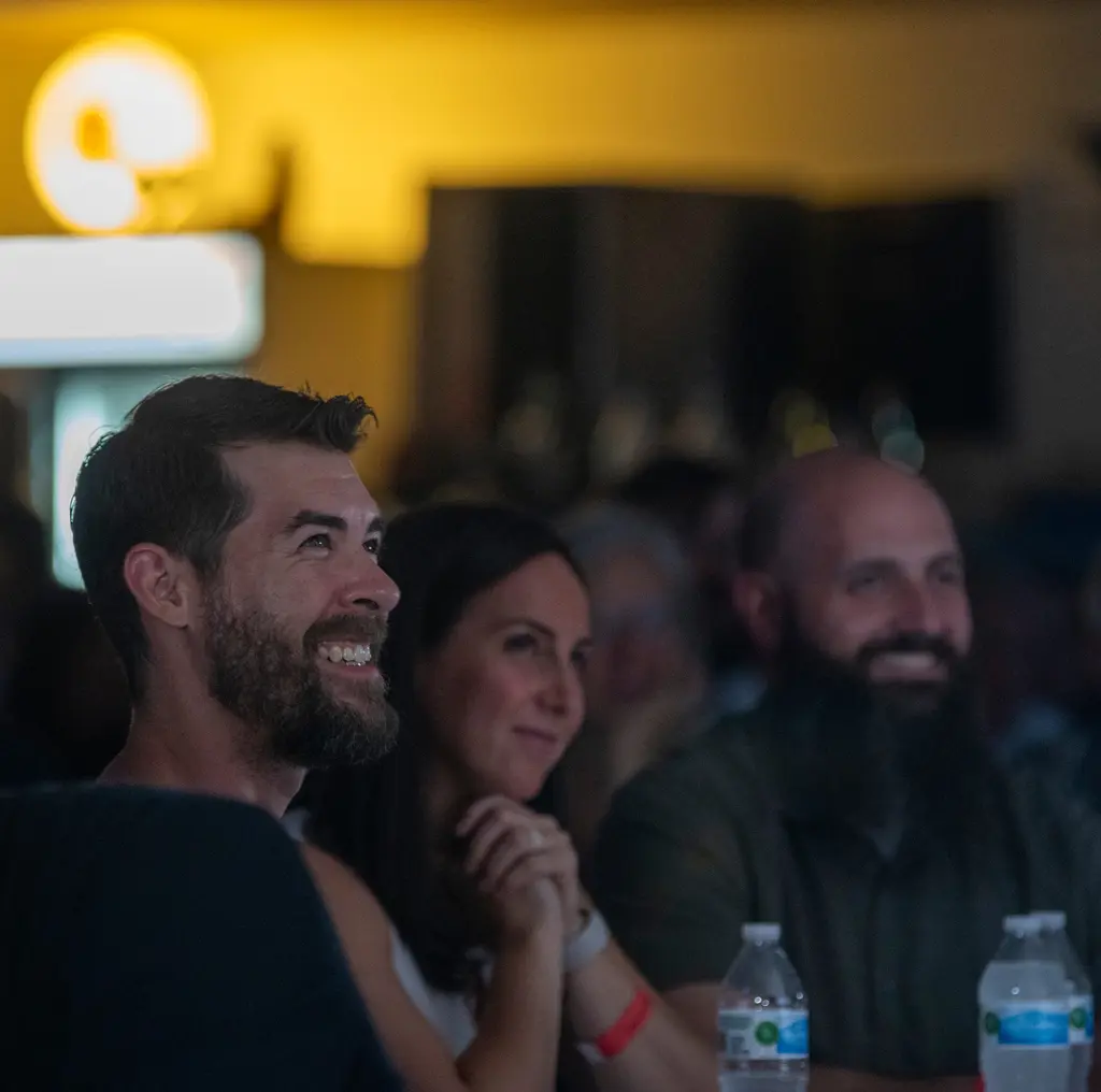 A bearded man in a black T-shirt smiles up toward the stage beside a woman with dark hair resting her chin on her hands, both lit by stage light in a dim room.