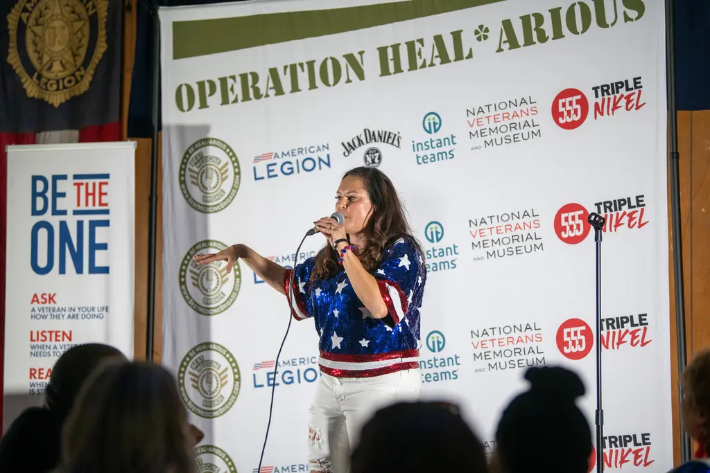 A comedian in a sequined stars-and-stripes top holds a microphone and stretches out one arm on a stage backed by an Operation Heal*Arious sponsor wall and a Be the One banner, with audience heads in the foreground.