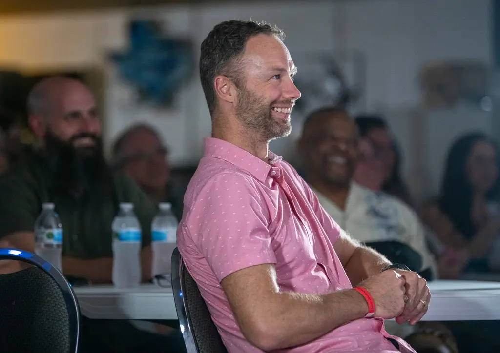 A bearded man in a pink short-sleeved shirt sits back in his chair and grins toward the stage, with other smiling guests blurred behind him.