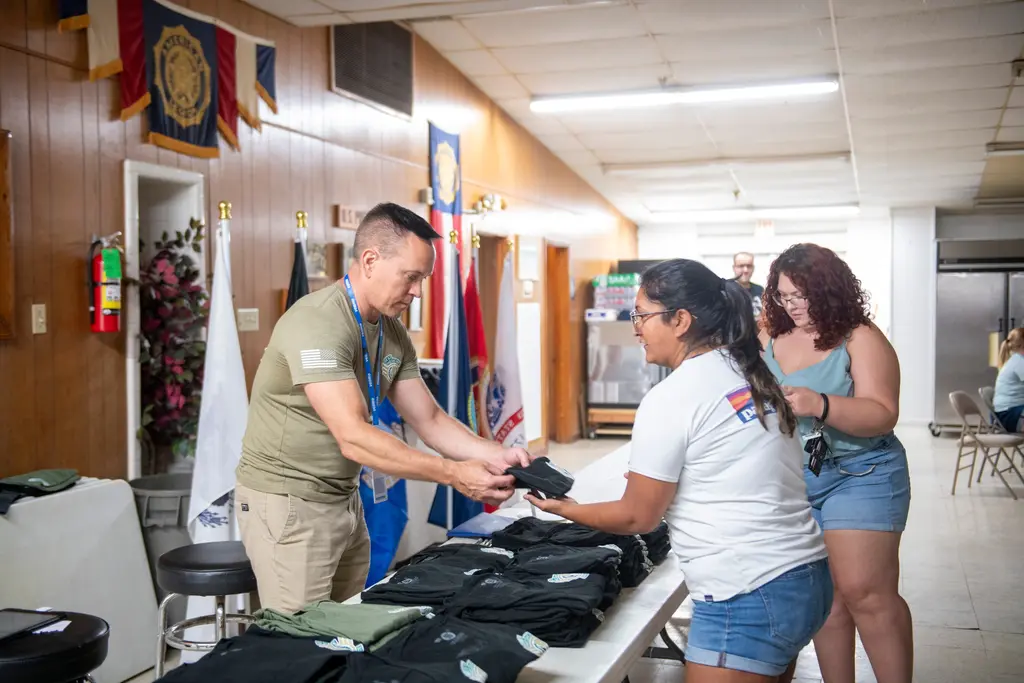 A man in an olive T-shirt and blue lanyard hands a folded black shirt across a table of stacked shirts to a woman in a white T-shirt as another woman looks on, in a wood-paneled hallway lined with flags.